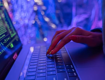 Person tippt auf Laptop-Tastatur; Monitor zeigt grüne Terminaltexte, blau-violette Lichtreflexe.