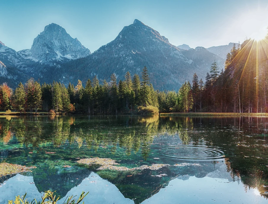 Bergsee, Uferbäume und Sonnenstrahlen rechts; klares Wasser spiegelt Berge und Himmel.